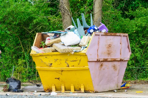 A large, rectangular skip with a yellow base and pinkish sides situated outdoors on a gravel surface near a wooded area. The skip is filled with a mixture of household waste, including white plastic bags, a blue plastic bucket, a purple container, and various shredded paper and cardboard. Some items, like a white cloth and a piece of foam or insulation, are visible among the clutter. The background features green foliage and tree trunks, indicating a rural or suburban setting. The skip is positioned close to a natural environment, with the presence of green leafy trees behind it, suggesting an area where garden clearance or outdoor rubbish removal services by Garden Clearance Archway may be needed. The overall scene conveys a typical rubbish disposal site, with the skip overflowing with mixed waste materials intended for collection or removal.