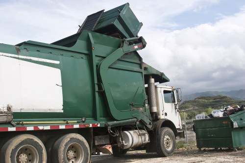 A large green waste collection truck is parked on an outdoor gravel area, with its rear compaction body raised to load rubbish. The truck's metal surface shows signs of wear and weathering, with visible textures of painted metal and hydraulic components. To the right of the truck, there is a green skip filled with mixed waste, including bags and loose debris. The scene is set against a backdrop of distant hilly terrain under a partly cloudy sky, suggesting an outdoor waste removal operation typical of horticultural or general rubbish clearance in a rural or suburban setting. The setting indicates professional waste disposal services often associated with garden clearance or rubbish removal in areas like [POSTCODE] or nearby regions, as operated by Garden Clearance Archway.