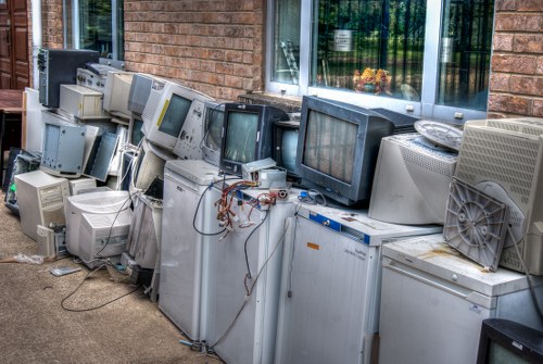 A row of discarded old computer monitors, CPUs, and electronic equipment lined up along a brick building exterior with a large window. The electronic devices vary in size, shape, and colour, predominantly in shades of grey, beige, and black, and appear to be placed directly on the pavement, suggesting a local waste clearance or electronic rubbish removal operation. Some devices show visible cables and wires, with a few monitors leaning or overlapping. The background includes a brick wall and a window reflecting greenery, indicating an outdoor environment possibly adjacent to a garden or residential property. The overall scene depicts an accumulation of electronic waste awaiting collection, representative of rubbish removal services offered by companies such as Garden Clearance Archway in the London or nearby areas. The image emphasizes the need for proper disposal of outdated electronic items as part of environmentally responsible waste management and garden clearance tasks.