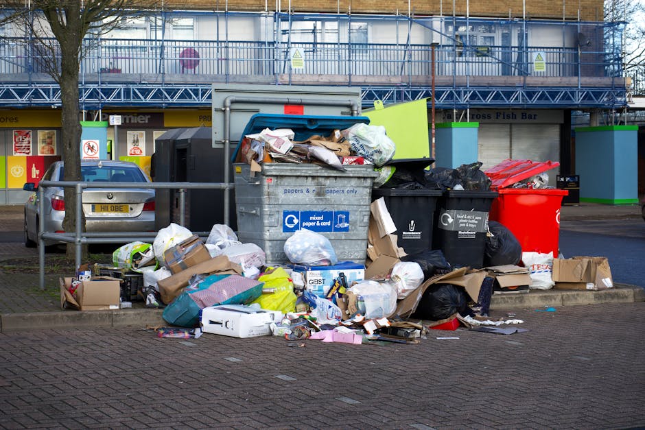 A large collection of overflowing waste and rubbish bags, cardboard boxes, paper and plastic packaging, and loose debris piled around a metal communal bin on a paved roadside area near parked vehicles