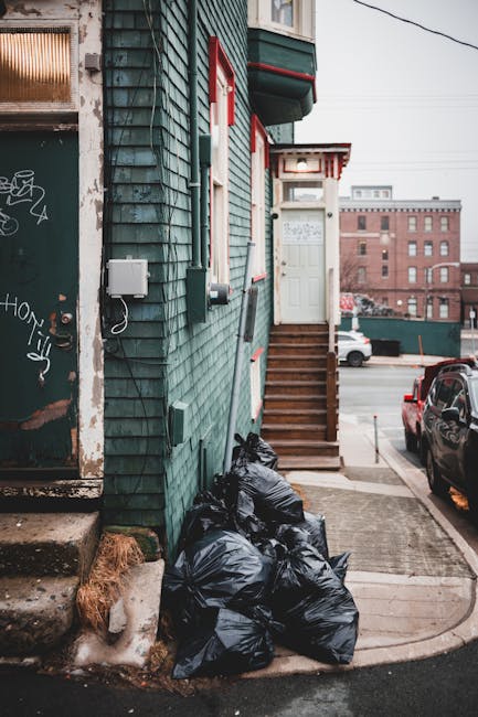 A large collection of overflowing waste and rubbish bags, cardboard boxes, paper and plastic packaging, and loose debris piled around a metal communal bin on a paved roadside area near parked vehicles